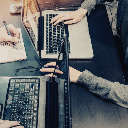 Close-up of 2 people working across a table from each other, each with a laptop in front of them
