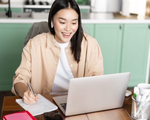 Young asian woman taking notes, working during video conference with laptop at home.